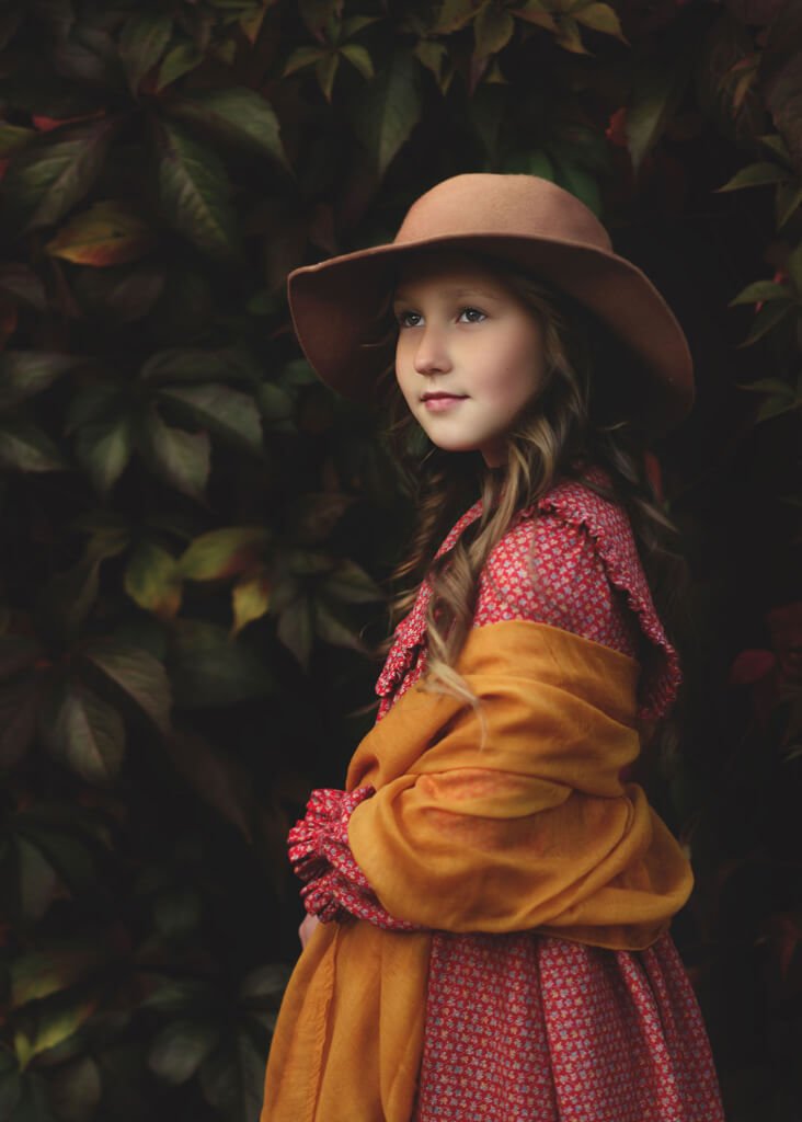 A young girl in a rustic hat and golden coat stands before a backdrop of autumn leaves, captured by a Nottingham family photographer.