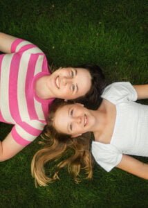 Two girls lying on green grass smiling during a playful photoshoot for kids.