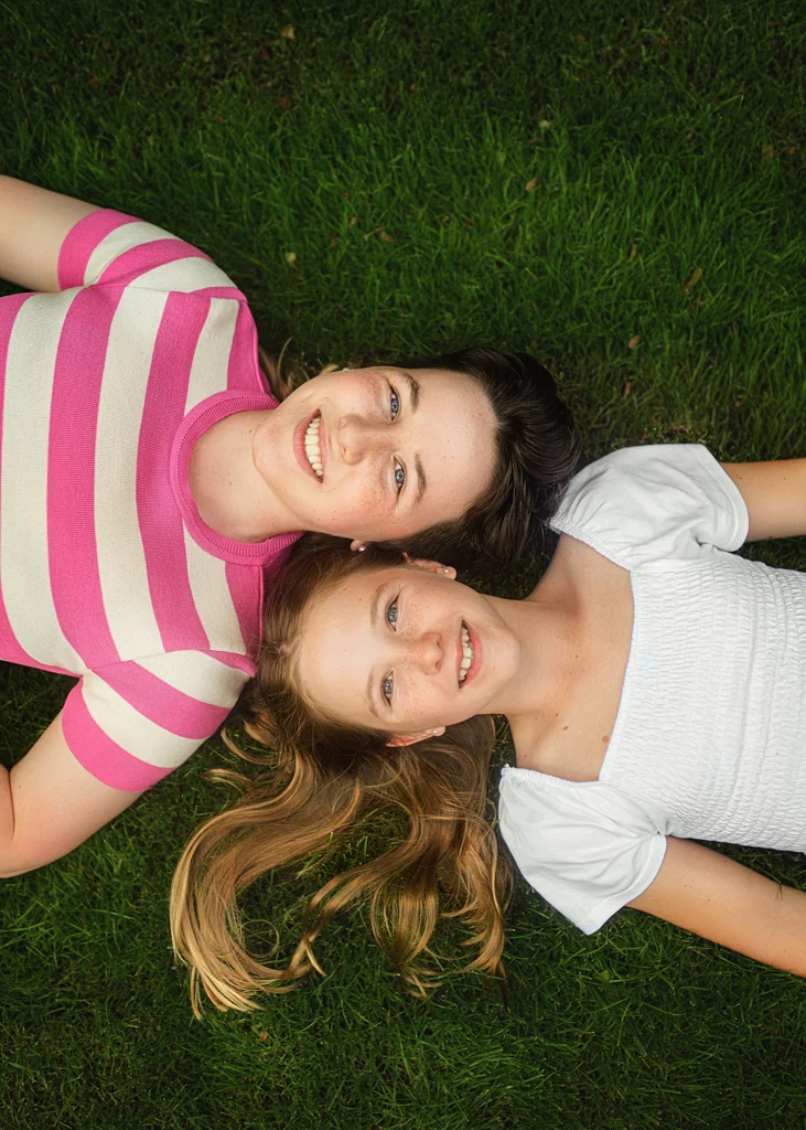 Two girls lying on green grass smiling during a playful photoshoot for kids.
