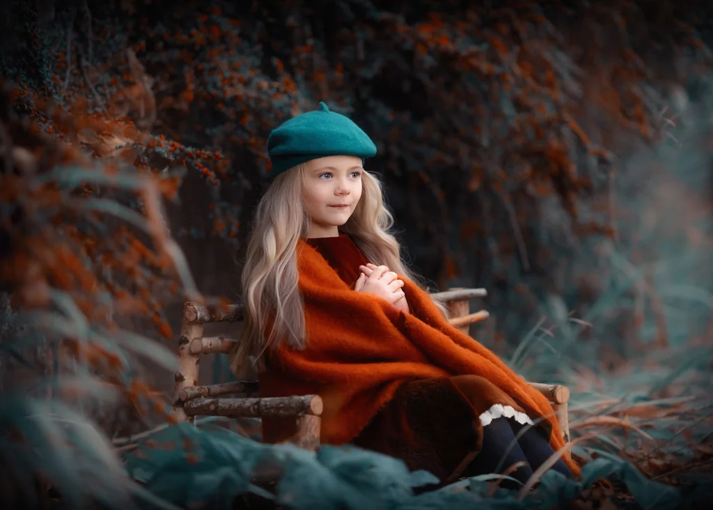 Young girl in a green beret and autumn cloak sitting on a wooden bench during a whimsical photoshoot for kids.