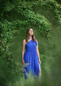 A contemplative woman in a vibrant blue dress stands amidst lush greenery, embodying the serene beauty of Nottingham.