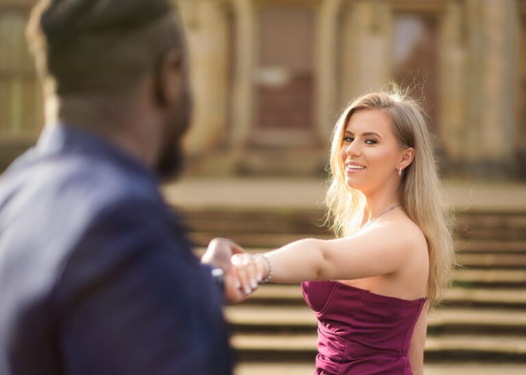 Engaged couple in a loving embrace, with the woman in a burgundy dress smiling over her shoulder at the camera, as her fiancé holds her hand, set against a grand architectural backdrop
