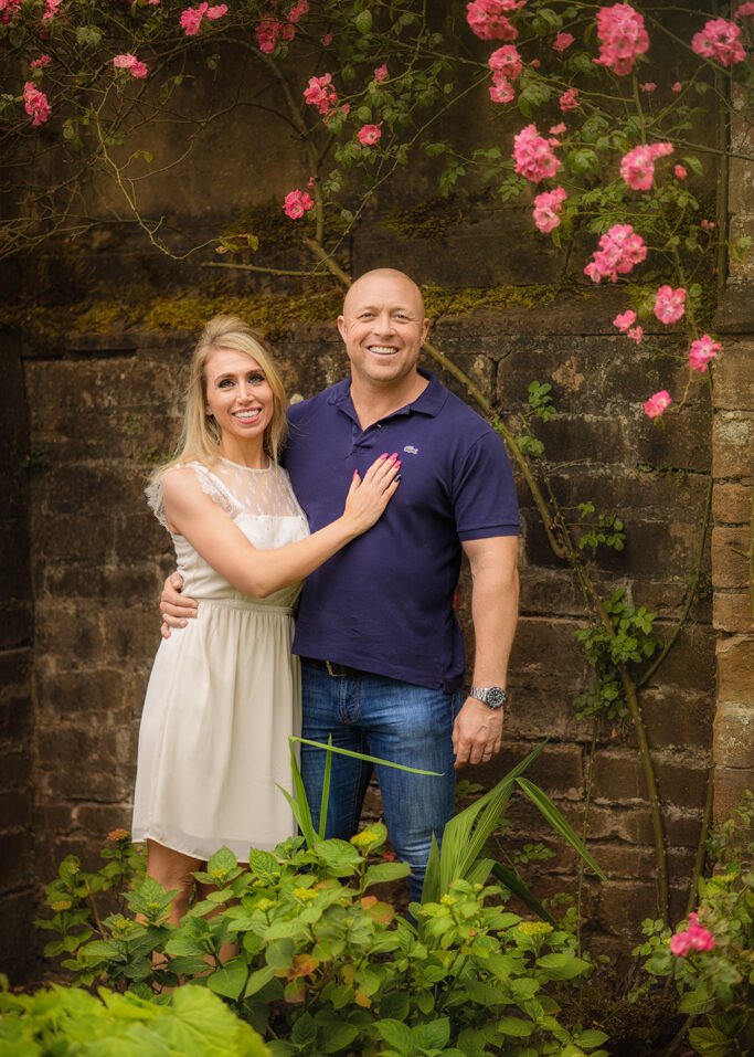 Joyful couple posing in front of a rustic stone wall adorned with blooming pink roses