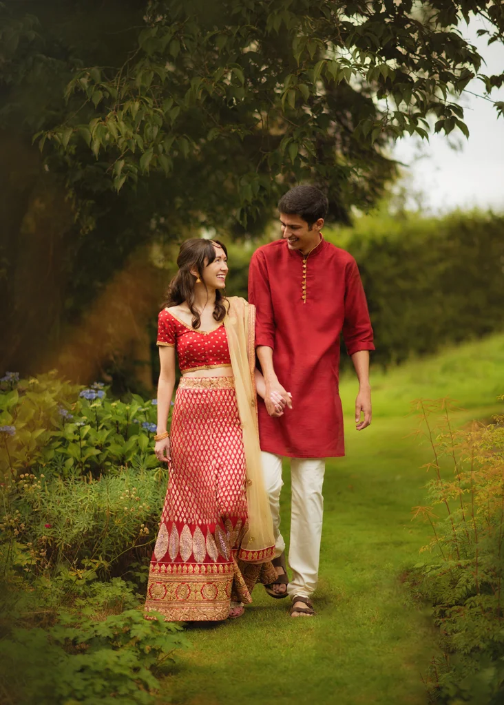 Couple dressed in traditional attire walking hand in hand during a pre-wedding photoshoot in a lush garden.
