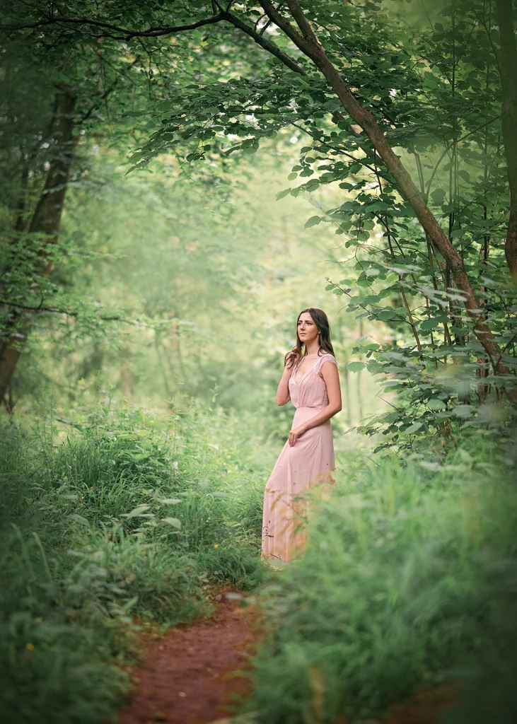 A pensive woman in a blush dress stands amidst a tranquil woodland path in Nottingham, reflecting the peace of her surroundings.