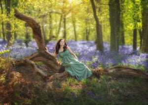 Glamour woman sitting on an old tree with a huge field of Bluebells in the background, portrait of a mum during family outdoor photoshoot in Nottingham Eastwood