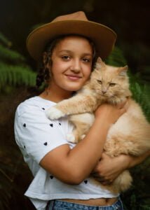 A young girl in a rustic hat shares a tender embrace with her fluffy ginger cat, a moment of pure affection captured amidst the greenery.