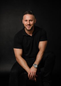 Business photography portrait of a confident man in a black polo shirt sitting against a dark background.