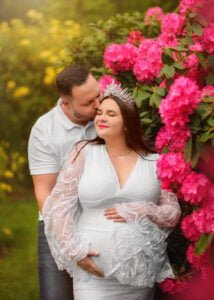 Surrounded by vibrant blooms, a loving partner kisses the forehead of the smiling mother-to-be adorned with a tiara, as they bask in the beauty of impending parenthood.