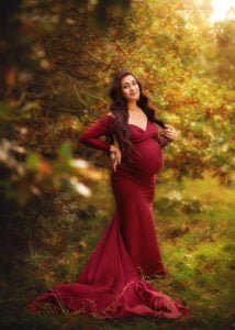 A radiant mother-to-be stands amidst autumn foliage, her flowing burgundy gown mirroring the natural palette of the maternity photography setting.