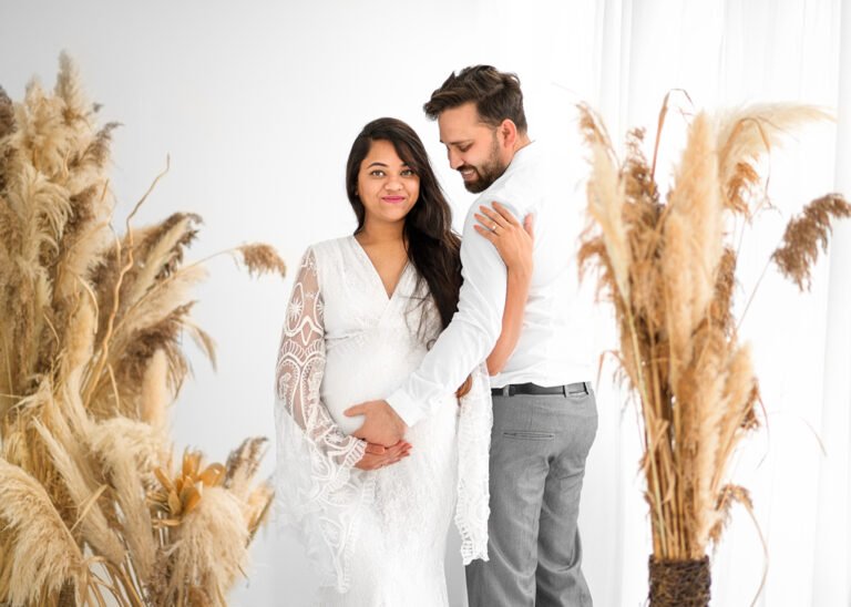 A couple stands in harmonious anticipation, with the expectant mother in a delicate white lace dress, cradled by her partner against a backdrop of golden pampas grass.