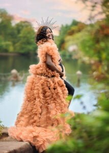 A radiant mother-to-be exudes happiness in a striking peach ruffle gown by the lakeside, her unique headdress a symbol of the transformative power of maternity.