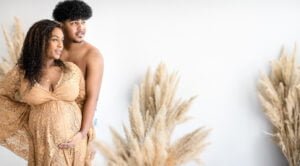 Expectant couple poses during a maternity session, with the mother-to-be in a beige lace dress in a natural light-filled studio.