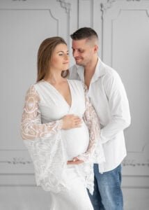 In an elegant white setting, a couple shares a quiet moment, the expectant mother in a lace-detailed dress and the father with a tender smile.