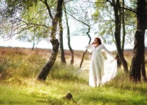 Red-haired mature woman in a flowing white dress standing gracefully in a sunlit meadow surrounded by trees.