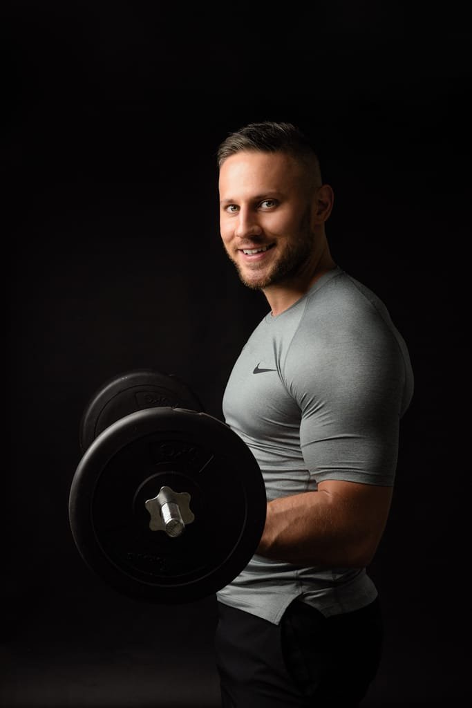 Business photography of a fit personal trainer lifting a dumbbell against a dark background.