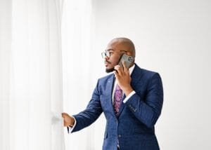 Professional men’s photoshoot featuring a confident man in a navy blue suit talking on the phone near a bright window.