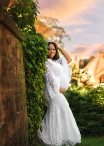 A serene expectant mother in a white lace dress stands crowned and radiant against an ivy-draped wall at golden hour, her joy unmistakable.