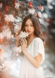 Young woman in a white dress holding delicate flowers under a blooming tree, ethereal professional portrait.