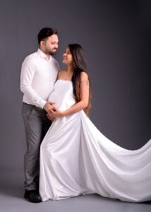 A loving couple stands intertwined, the man in a crisp white shirt and the woman in a flowing white maternity dress, both smiling tenderly at each other against a soft grey backdrop.
