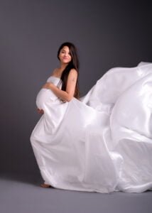 A beaming mother-to-be in a flowing white dress cradles her baby bump, her joy tangible in this ethereal maternity photography portrait.