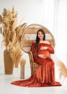 A glowing mother-to-be seated elegantly in a peacock chair, her rust velvet gown flowing beautifully, surrounded by the natural textures of pampas grass.