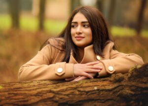 Reflective birthday girl in a cozy camel coat leaning on an ancient tree during an outdoor photoshoot.