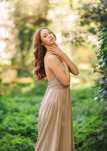 Elegant woman in a beige dress stands amidst a sunlit forest for her outdoor birthday photoshoot.