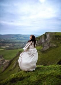 Elegant lady in a flowing white gown stands atop a lush green cliff, celebrating with an outdoor birthday photoshoot.