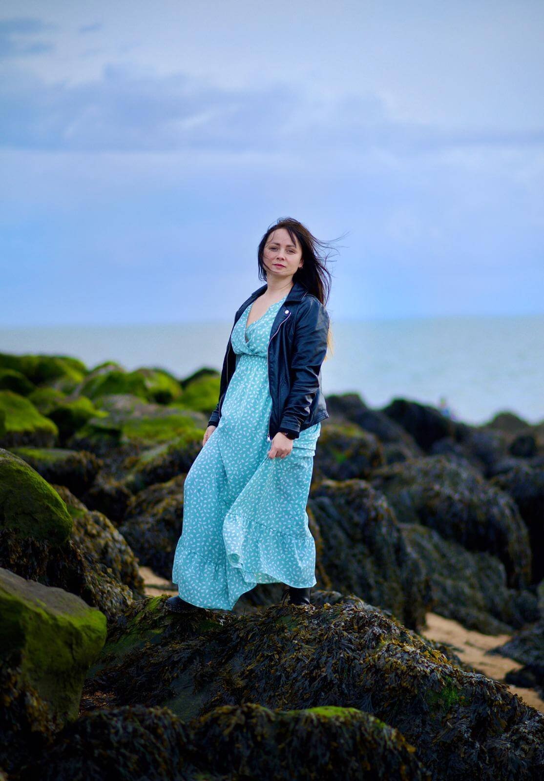 Woman in Teal Patterned Dress and Black Leather Jacket by the Sea