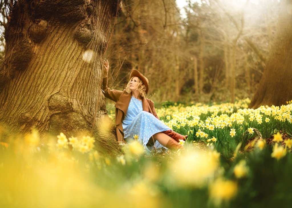 A thoughtful woman in a flowing blue dress and stylish hat sits by an ancient tree, her gaze uplifted to the forest canopy above, while the ground around her is carpeted with the bright cheer of daffodils.