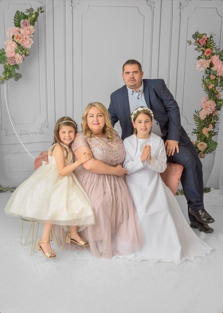 A family gathers closely, their joy palpable, as they celebrate a child’s first holy communion, with an elegant floral backdrop enhancing the ceremonial ambience.