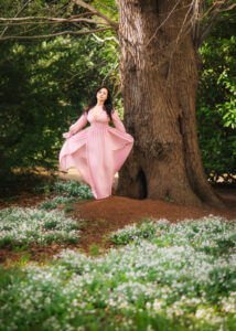 A woman in a flowing pink gown stands with a graceful poise in a serene woodland, her dress billowing softly, as she looks off into the distance, a vision of elegance and serenity among the blossoming flowers.