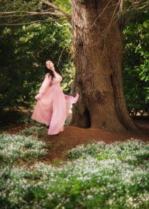 A woman in a flowing pink dress closes her eyes and lifts her face towards the sky, her posture and expression full of grace and ease, as if absorbing the forest's tranquil energy amidst a blooming white flowerbed.