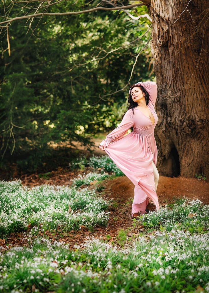 A woman in a flowing pink dress embodies grace and movement as she glides through a tranquil woodland setting, her pose expressive of freedom and self-discovery amidst the springtime bloom.