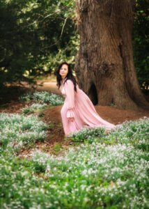 A woman in a long pink dress kneels gracefully among a field of delicate white flowers, her gaze thoughtful and inviting, as she connects with the timeless beauty of the natural world.