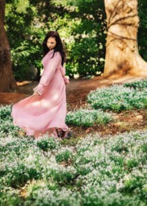 A woman in a flowing pink dress takes a leisurely stroll through a white flower-laden path in the woods, her movement full of grace and her expression one of contentedness, in tune with the forest's tranquil beauty.