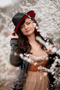 A woman with a serene expression gently adjusts her stylish red and black hat, surrounded by the delicate white blossoms of early spring, symbolizing a peaceful harmony with the budding nature around her.