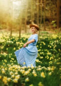 A joyful woman in a blue floral dress and chic tan hat stands amidst a vibrant field of yellow daffodils, her playful twirl and radiant smile capturing the essence of spring's exuberance.