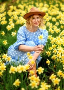 A woman with a tender smile, wearing a blue dress and a tan hat, sits gracefully among vibrant yellow daffodils, holding a single flower as a symbol of her connection to nature and her own blossoming self.