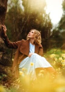 Nottingham Female Portrait Photography – A woman in soft blue and brown tones gazes up at a tree, bathed in golden light, embracing nature and self-discovery.