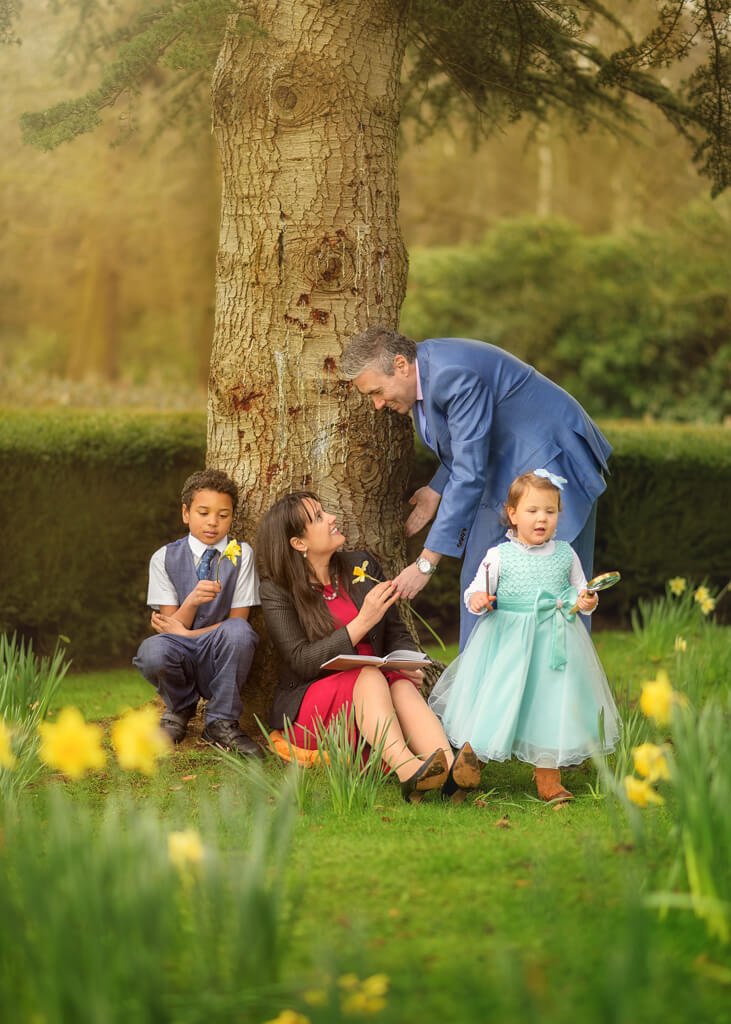Family sitting under the tree during a spring outdoor photoshoot at Elvaston Castle. Dad handing a daffodil flower to mum - Nottingham Family Photographer.