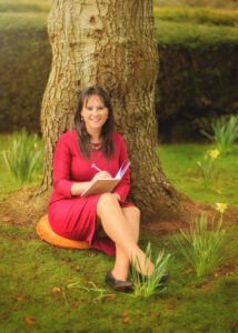Portrait of a mum writing in her diary and sitting in a red dress under a tree at Elvaston Castle Park during a professional family outdoor photoshoot in spring.