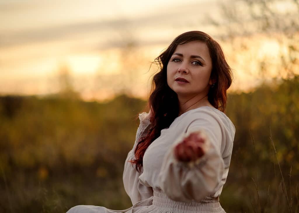 Woman in a serene white dress extends a hand, contemplating the amber sunset during her outdoor birthday photoshoot in the countryside.