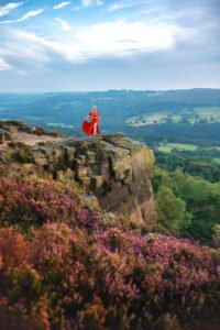 Woman in flowing red dress standing atop a majestic cliff, surrounded by wild heather, with a panoramic view of the valley below.