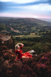 A woman in a red dress reclines gracefully on a cliff at Mother Cap, gazing towards the sunset horizon.