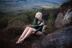A contemplative woman in an elegant green bodysuit sits among the heather at Mother Cap, her gaze reflective and serene.