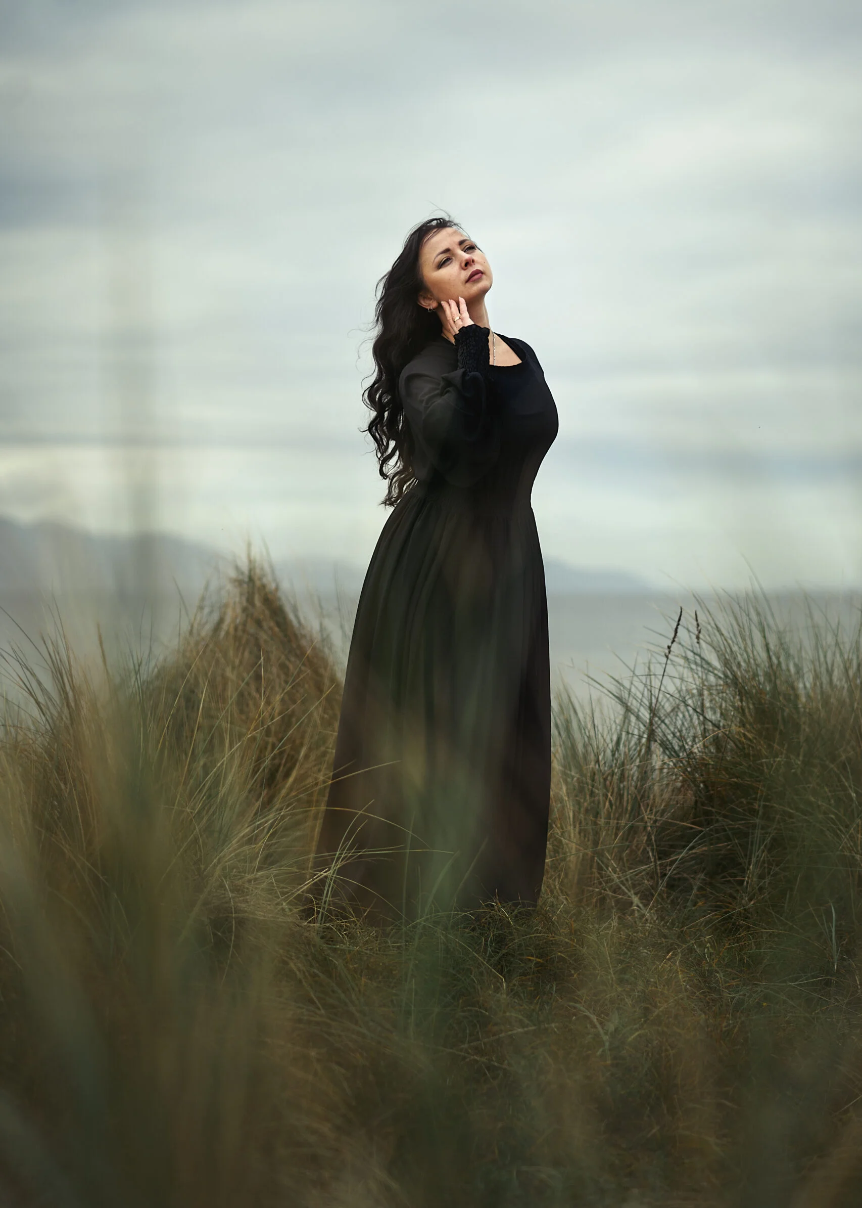 Mature woman in a long black dress standing amidst tall grass, exuding quiet confidence against a dramatic sky.