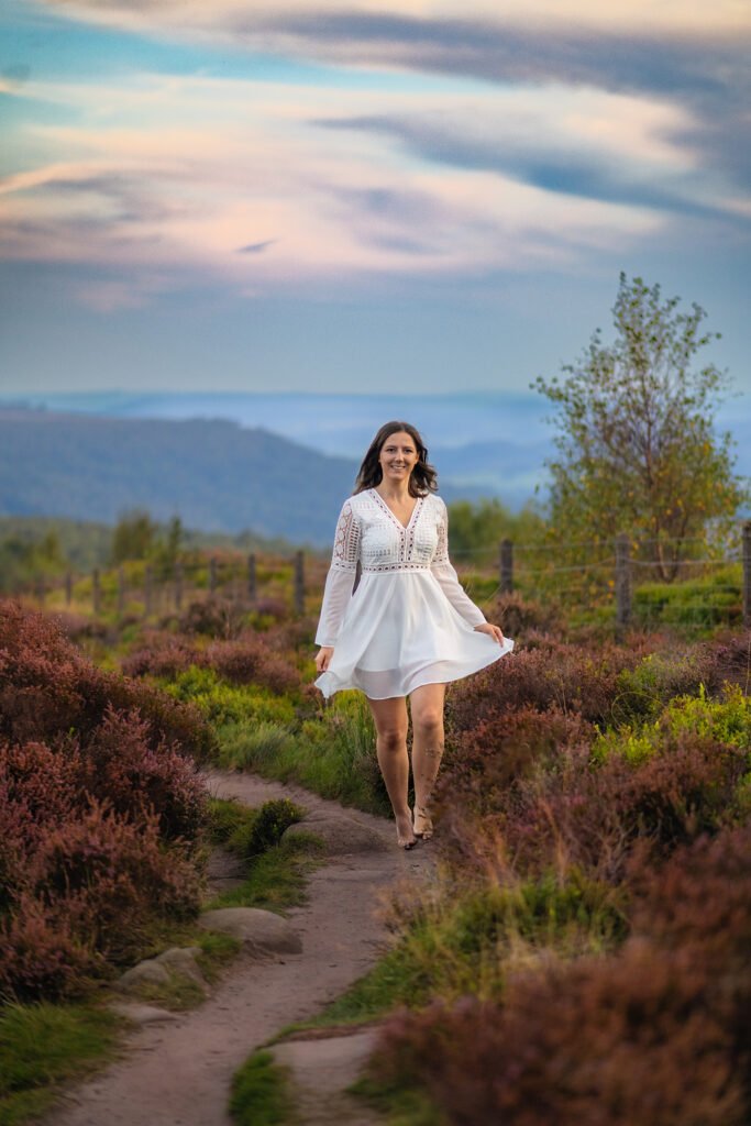 Joyful woman in a white dress walking along a path at Mother Cap, surrounded by heather, embodying freedom and grace.