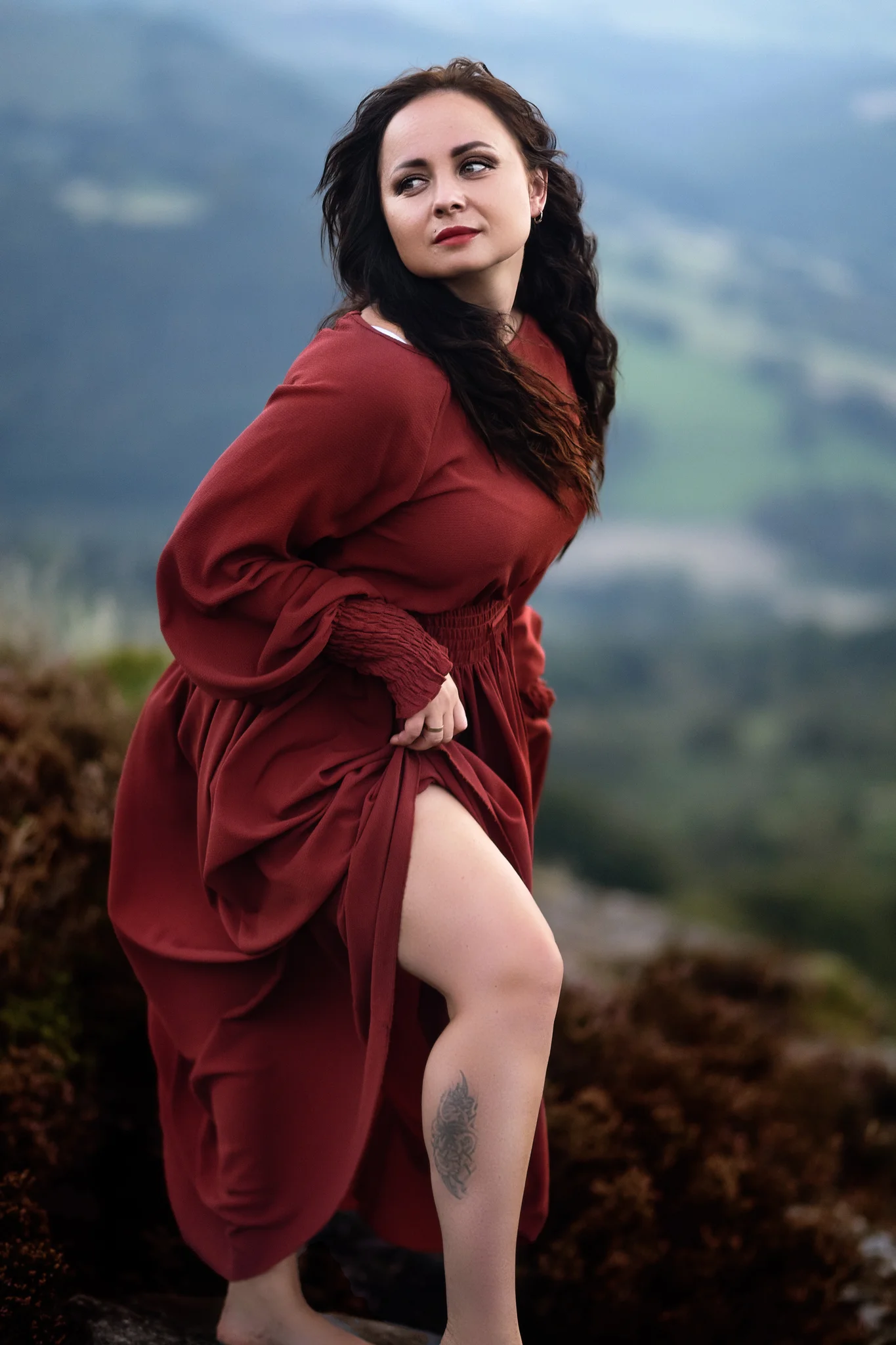 A thoughtful woman in a russet dress stands against the misty backdrop of Mother Cap, her tattoo a whisper of her story.